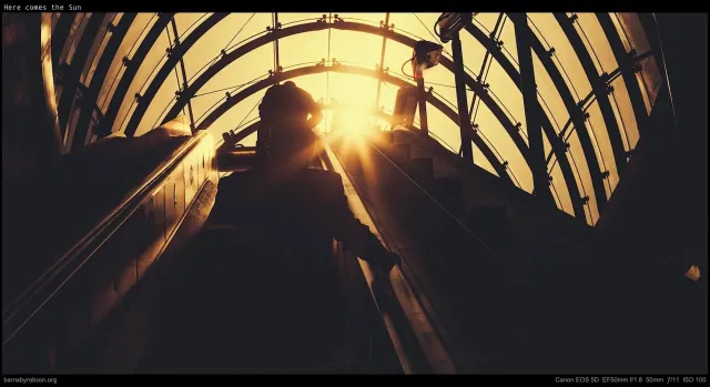 Commuters descending into a London station as low sun bursts through the ironwork, black and white.