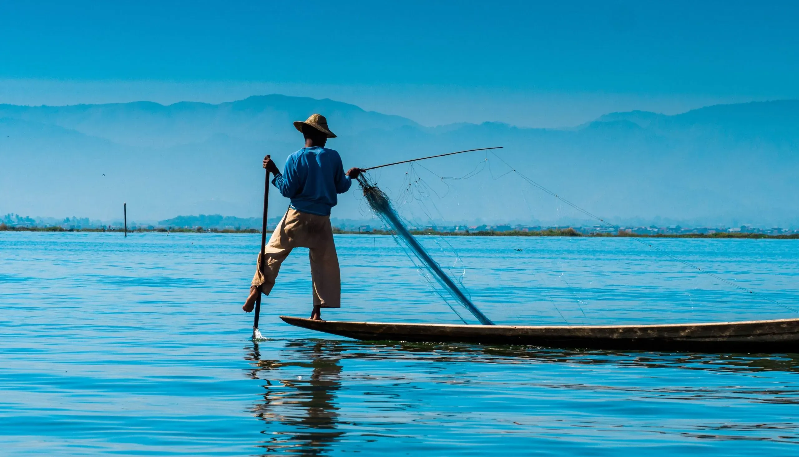 half An Intha fisherman rowing one-legged on Inle Lake, net in hand against blue haze