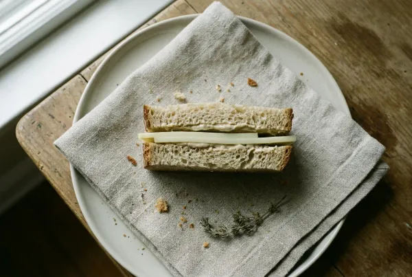 A simple handmade sandwich cut in half on a linen napkin and pale plate in soft window light, its layers on show — a nod to the human-AI sandwich: you frame the task, the machine fills the middle, you finish it.