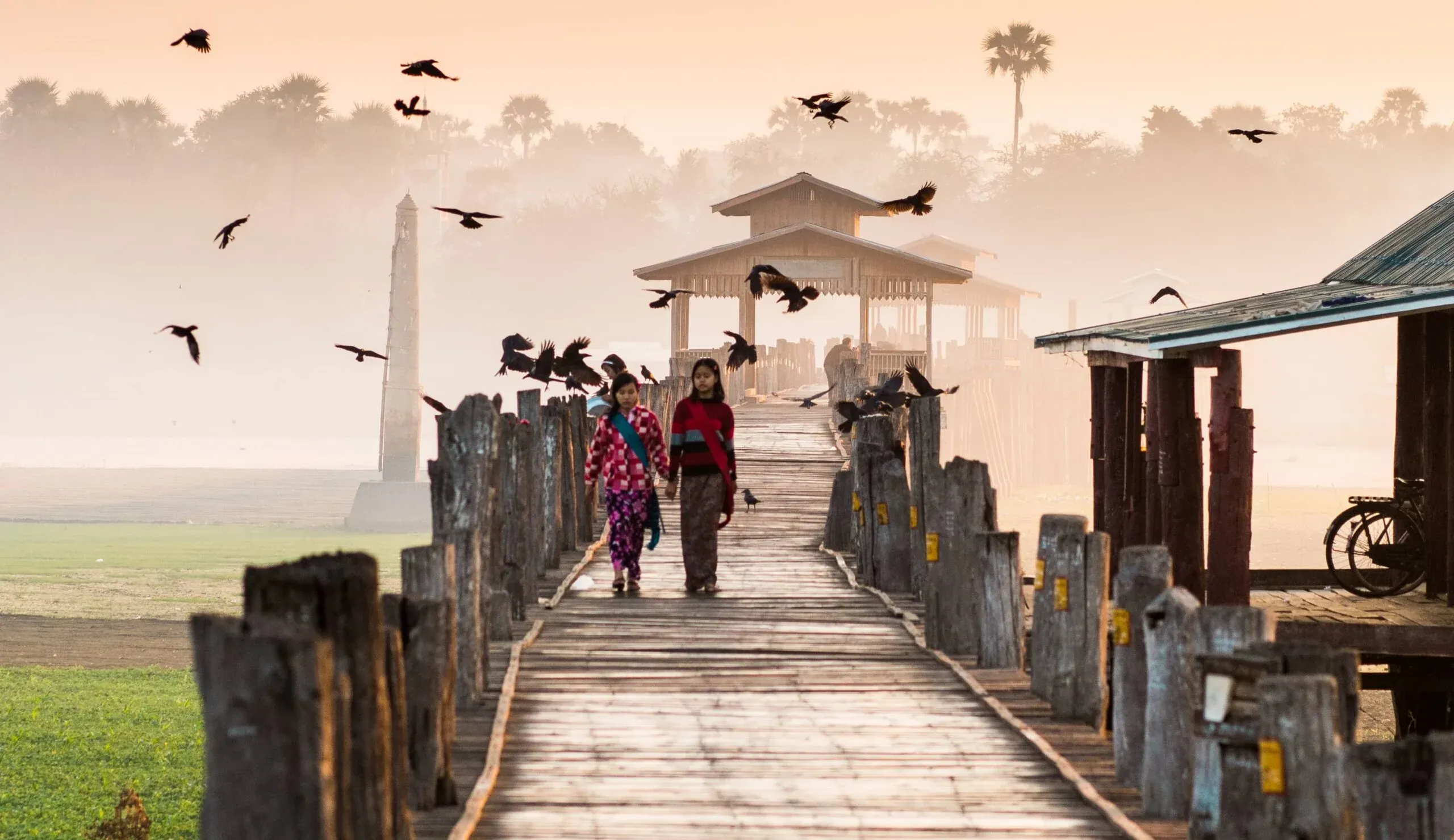 wide U Bein Bridge at dawn: two girls on the teak walkway, crows wheeling through the mist